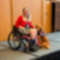 {image description: Allee, a white woman with glasses, blonde hair, facial piercings, and tattoos, is seated in her wheelchair on a stage with her Service Dog, Persie, next to her. Persie is a red, golden retriever with a hot pink service dog vest.}