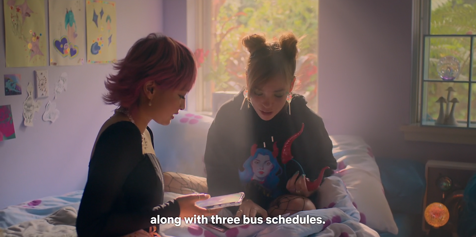 Quinni and sasha sit on a bed in a colorful room, Sasha holding a phone. Quinni reads from paper on the bed. Subtitles read: "along with three bus schedules."