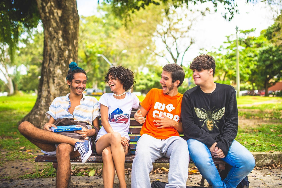 4 jovens adolescentes sentados num banco de jardim. À esquerda, um rapaz de sapatos allstar, camiseta de botão aberta e cabelo apanhado com tons de azul. A seguir, uma menina de roupa branca olha pra ele e ri. Sandro vem depois, com camiseta laranja escrita "rir cura" rindo e também olhando para o primeiro. O quarto é um jovem de calça jeans e camisa de manga comprida preta, com um sorriso sem mostrar os dentes.