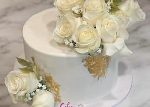 A simple white cake is adorned with white roses and gold leaf accents, creating an elegant and modern design.