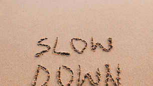 A decorative image that has the term "slow down" written in the sand at a beach with waves.