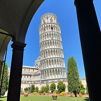 The Leaning Tower of Pisa seen in the distance from beneath a shaded archway, which frames the tower and sky.
