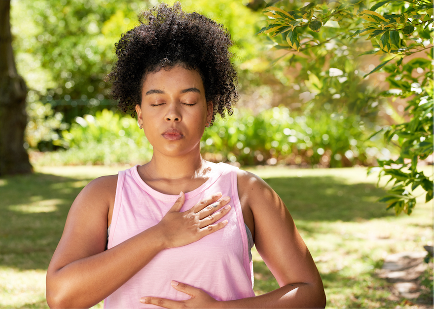 Woman in pink tank top meditates with eyes closed and hand on chest in a sunny green park.