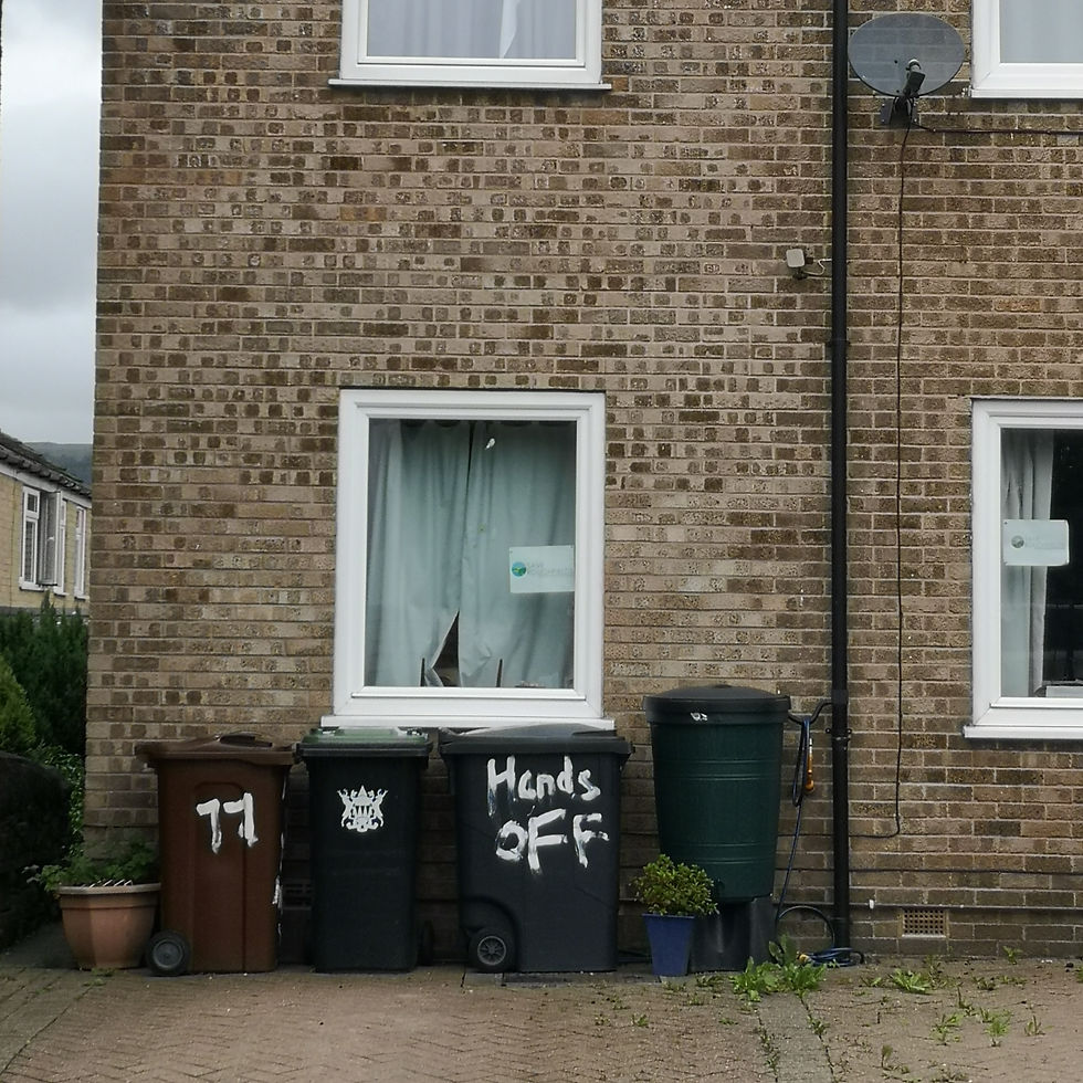 Brick house facade with two windows, curtains, and a satellite dish. Three trash bins, one with "Hands OFF" graffiti, stand on the driveway.