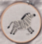 Embroidery of a running zebra on beige fabric in a hoop, featuring black stripes and text "MN" in the corner, against a soft textured background.