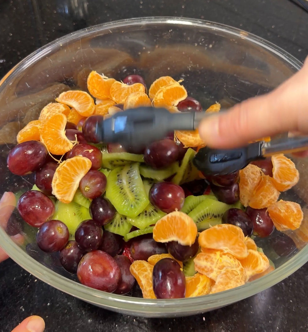 A hand uses black tongs to mix a colorful fruit salad with grapes, kiwi, and orange slices in a clear bowl on a dark countertop.