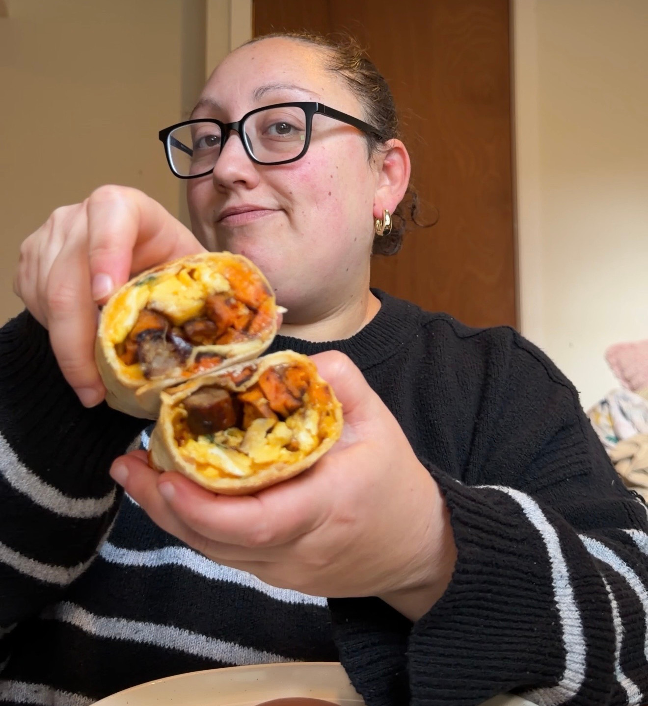 Woman in glasses holds up a cut burrito over a plate with sauce in a cozy home kitchen, looking pleased.