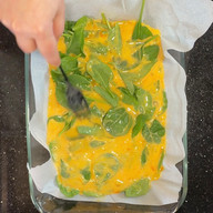 hand using form to push spinach around in eggs that are poured into a glass pan