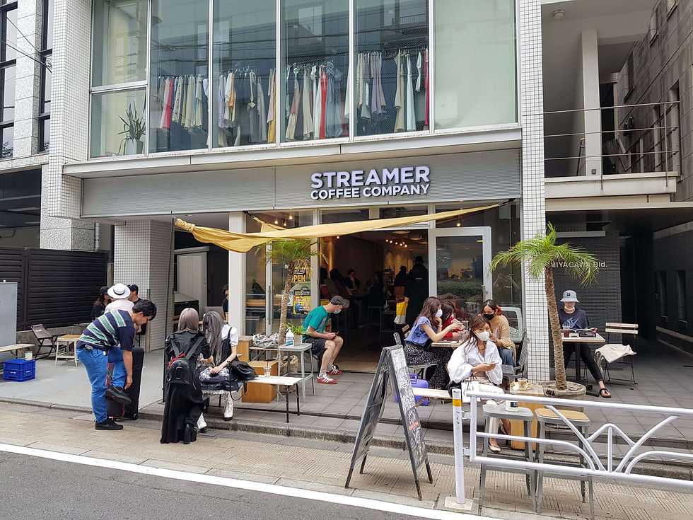 People relax outside Streamer Coffee Company, sitting at tables with drinks. A small palm tree decorates the entrance. Casual, urban setting.