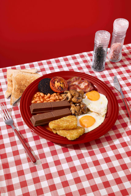 Playful full English breakfast scene with fried eggs, toast, mushrooms, tomato, hash browns, baked beans, and a KitKat Chunky chocolate bar on a red checked tablecloth. Commercial breakfast food photography by Richard Jarrett, London.