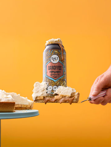 Commercial drinks photography of SALT Banoffee PUDD stout balanced on a banoffee dessert with whipped cream and caramel, styled on a bold yellow studio background by advertising photographer Richard Jarrett, London.
