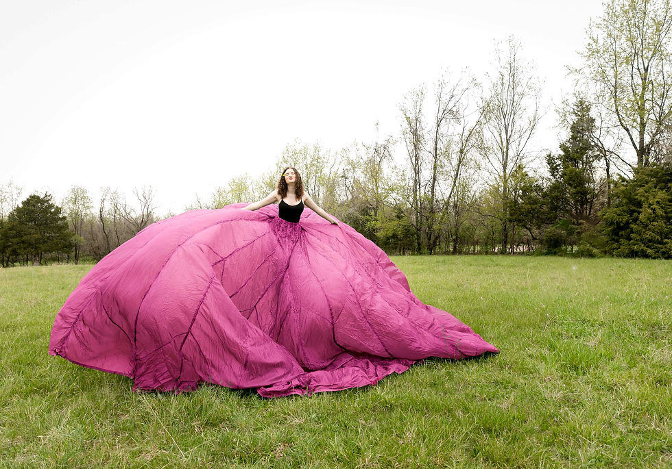 Woman in a flowing pink dress stands in a grassy field. Her arms are spread, and trees are in the background. The mood is serene.