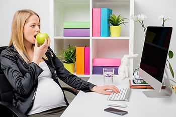 A white pregnant woman with blond hair eating an apple while sitting at a desk. 