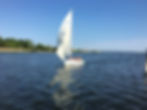Sailboat with people in life vests glides on a blue lake, under a clear sky. Lush green trees line the distant shore.