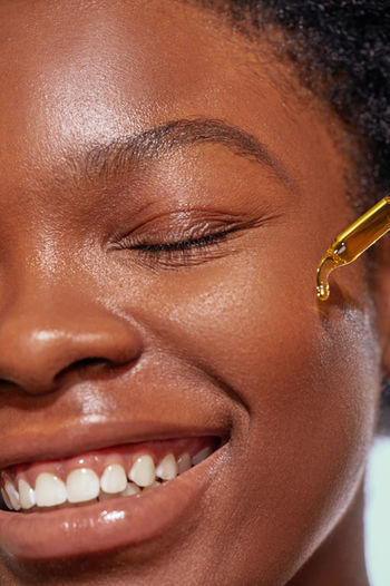 Super close up half face shot of natural makeup on black female model, smiling with eyes closed, dropping oil onto her face.