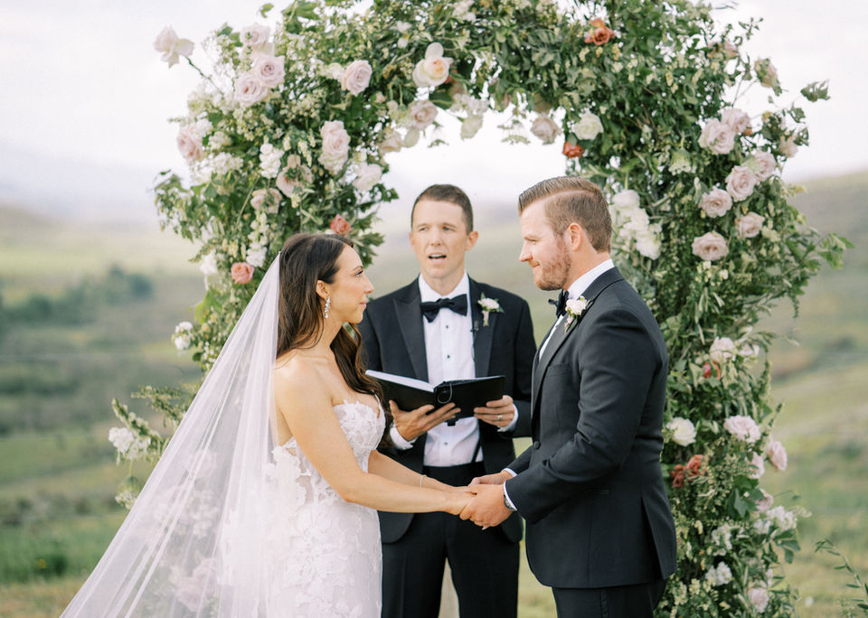 A couple look deeply into each others eyes in front of a floral covered arch as they say I do at a ranch wedding venue in Colorado.