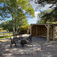 Wooden pent gazebo next to a stone patio with fire pit and chairs.