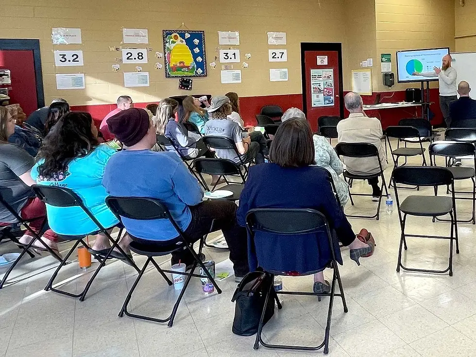 Several people sit in folding chairs while listening to a speaker at a Blythe Oldfield Community Association meeting.