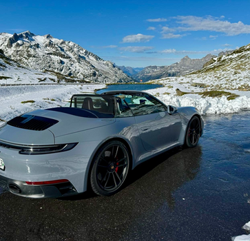 Porsche 911 Cabriolet on a mountain road, surrounded by fresh snow under a clear blue alpine sky.