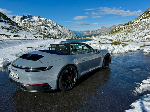 Porsche 911 Cabriolet on a mountain road, surrounded by fresh snow under a clear blue alpine sky.