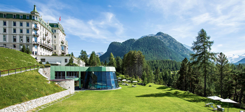 Exterior of the Grand Hotel Kronenhof in Pontresina with manicured lawn, loungers and forested mountains in the background.