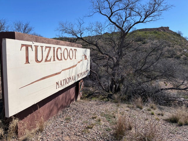 Sign reading "Tuzigoot National Monument" with barren trees and rocky terrain in the background under a clear blue sky.