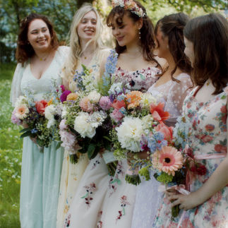 The bride stands with her bridesmaids, all holding vibrant bouquets of wildflowers, smiling together in a meadow setting.
