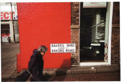 Disposable camera photo of an elderly woman walking past a bright red wall with a street sign reading “Bakers Yard leading to Bakers Road.