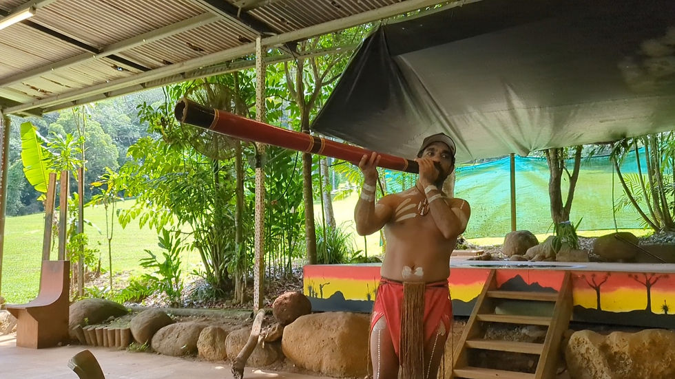 An Aboriginal man plays a long didgeridoo under a tin-roof shelter in a lush tropical setting, wearing body paint and red cloth.