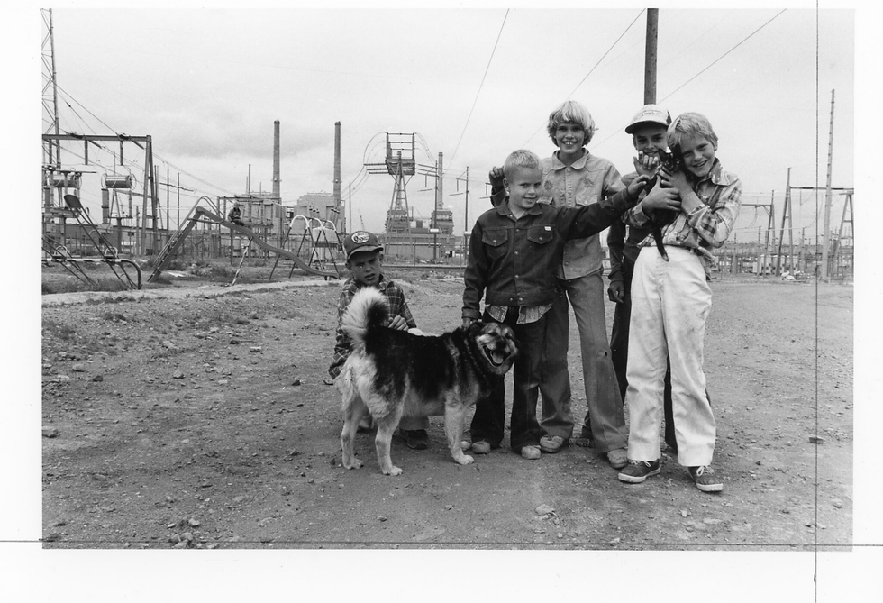 Five smiling children with a dog on a dirt road in front of power lines and smokestacks at an industrial site.