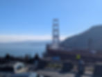 People enjoy the view of the Golden Gate Bridge, with cars crossing. Clear blue sky and hills in the background create a serene scene.