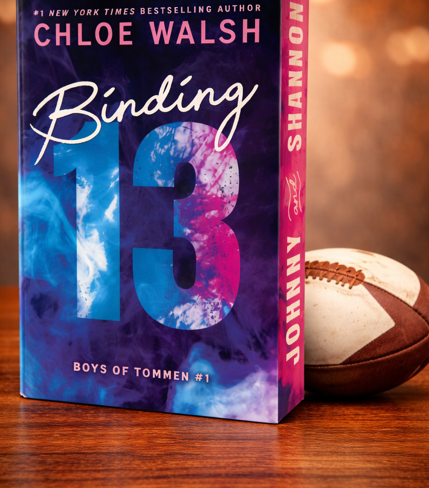 Book titled "Binding 13" by Chloe Walsh, with colorful blue and pink cover, on wooden table beside a football. Warm bokeh background.