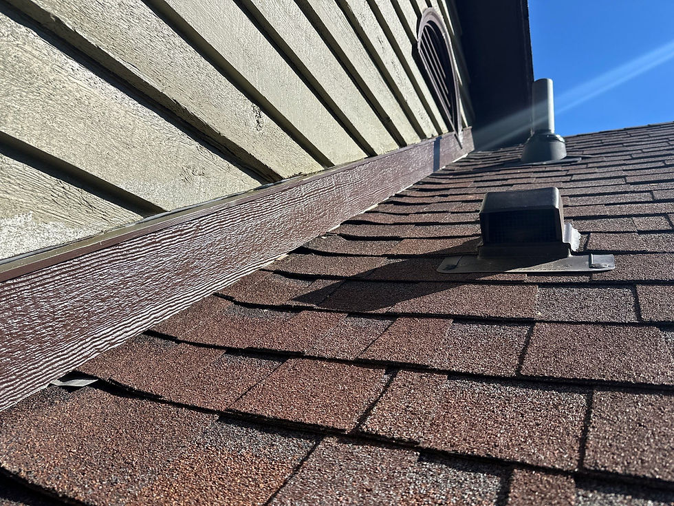 Close-up of a residential roof featuring brown architectural shingles, a black box-style roof vent, and brown wood-grain trim installed where the roofline meets light green lap siding