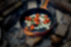 A heavy duty skillet cooking over a campfire surrounded by logs.