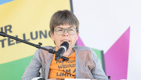 Eine Frau mit Brille, orangenem T-Shirt und grauer Strickjacke hält einen Zettel in den Händen. Ein schwarzes Mikrofon ist an einem Mikrofonständer zu ihrem Mund gerichtet. Im Hintergrund ein Plakat mit blauen, gelben, grünen und pinken Elementen. Text: UR TELLUNG in grossen Buchstaben.