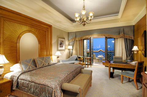 Luxurious guest room at Emirates Palace, Abu Dhabi, featuring gold-accented furnishings, marble flooring, recessed ceiling lighting, a classic chandelier, and floor-to-ceiling windows opening onto a waterfront balcony at night