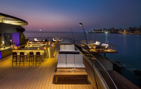 Nighttime view from a high deck overlooking an outdoor bar area and tranquil body of water, with ambient lighting and distant city lights creating a luxurious, atmospheric setting