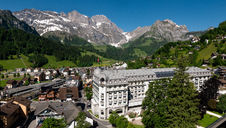 Scenic view of the Kempinski Palace Engelberg hotel in the Swiss Alps, surrounded by snow-capped mountains, lush green slopes, and a clear blue sky