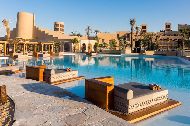 Bright, sunny day view of the large, blue swimming pool at Terra Solis in Dubai, with modern lounge beds, palm trees, and traditional earthen-toned buildings surrounding the area.