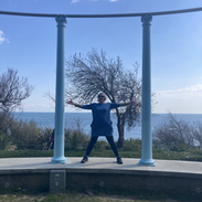 Woman stands between two roman style pillars, arms outstretched, sea behind her. 