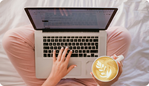 mulher sentada na cama, mexendo em seu notebook e tomando um café