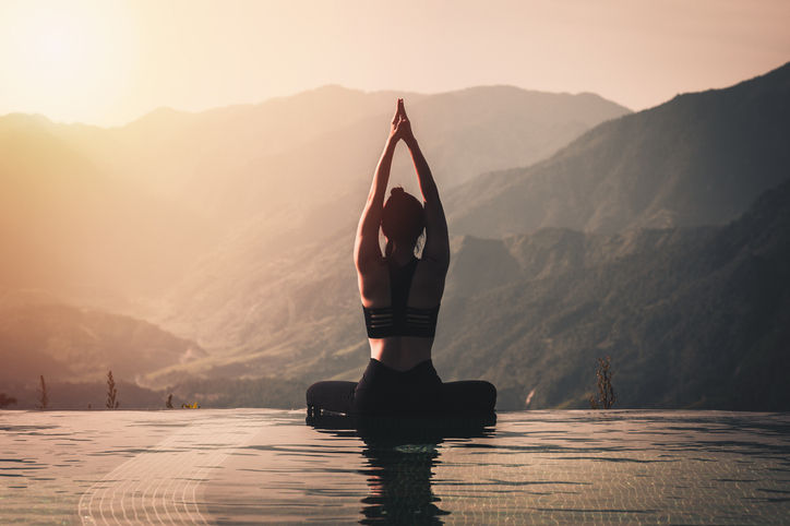 A serene, back-view composition of a person meditating in a seated yoga pose at the edge of an infinity pool. Their hands are raised and pressed together above their head in a prayer position. The calm water of the pool perfectly reflects the golden, hazy sunlight. In the background, layers of massive, misty mountain ridges stretch across the horizon under a soft, warm sky.