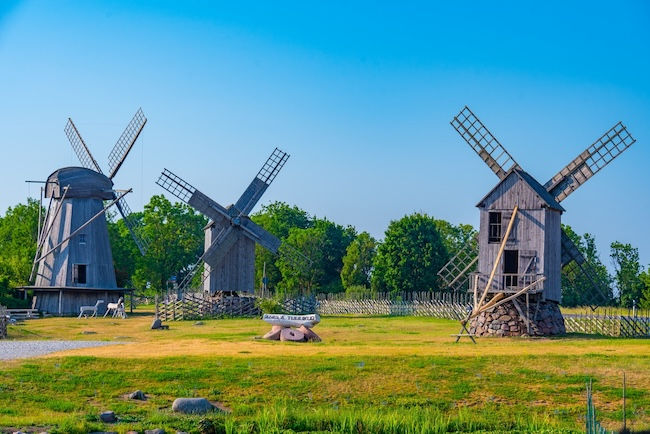 A row of three traditional wooden windmills stands in a grassy field at the Angla Windmill Mount in Saaremaa, Estonia. The windmills vary in design, with two featuring classic wooden trestles and four large sails, set against a backdrop of green trees and a clear, vibrant blue sky. A small wooden sign sits in the foreground.