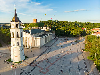 Aerial view of Cathedral Square in Vilnius under a bright blue sky. The massive white neoclassical Vilnius Cathedral and its iconic detached bell tower dominate the foreground, casting long shadows across the expansive stone plaza. In the distance, hot air balloons float above the green slopes of Gediminas' Hill.