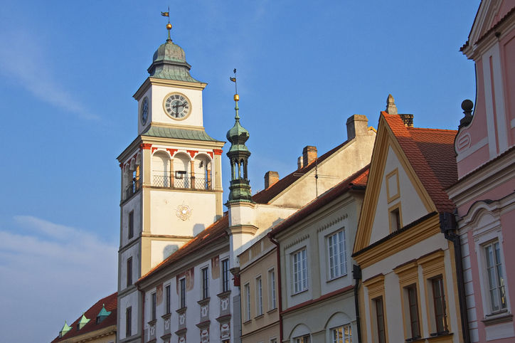 A photo of the Old Town Hall in Třeboň, Czech Republic, featuring its prominent square tower against a clear blue sky. The tower, which has a green cupola and a visible clock face near the top, is painted white with a red and green trim. It is attached to a row of historic buildings with steep, red-tiled roofs and ornate gables, including some painted in soft yellow and pale pink hues. The image is taken from a low angle, emphasizing the height of the architecture.