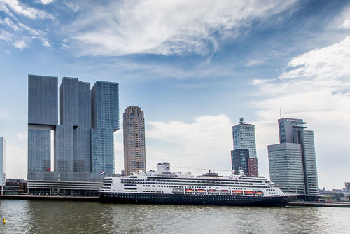 A large black and white cruise ship is docked in a body of water, with the modern skyline of Rotterdam, Netherlands, visible behind it under a cloudy sky. On the left, the "De Rotterdam" building, a striking complex of three connected towers, dominates the cityscape. Other contemporary skyscrapers and office buildings also rise from the waterfront.
