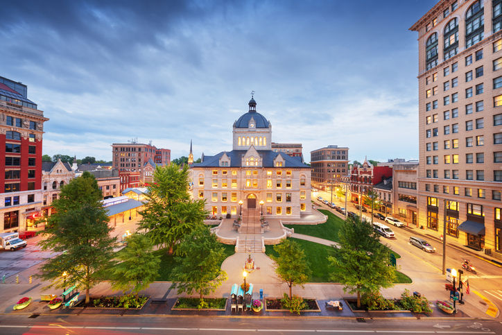 A wide dusk shot of the old Fayette County Courthouse in Lexington, Kentucky. The stone building is glowing with warm light against a blue twilight sky, centered among city streets, modern buildings, and lush green trees.