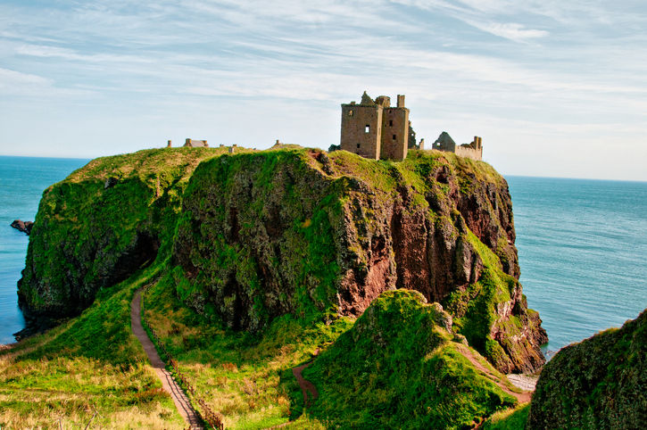 A breathtaking landscape of a ruined medieval fortress perched atop a massive, sheer-faced sea cliff. The cliff is covered in vibrant green grass and surrounded by the deep blue water of the North Sea. A narrow dirt path winds along the edge of the mainland leading toward the castle.