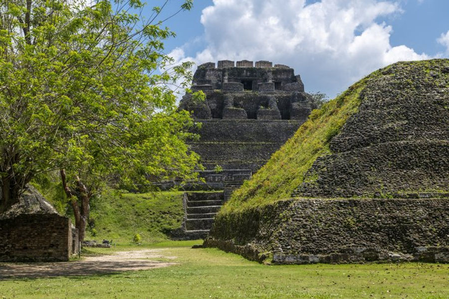 Ancient stone Maya pyramid structure at Xunantunich surrounded by grassy lawns and lush green trees under a partly cloudy sky