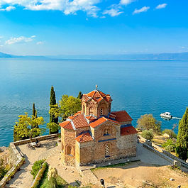 A high-angle view of the historic Byzantine-style Church of St. John at Kaneo situated on a cliffside promontory above Lake Ohrid in North Macedonia. The stone church features terracotta-tiled roofs and an octagonal dome, surrounded by cypress trees, green foliage, and stone pathways, set against the vast, deep blue expanse of the lake and distant mountains under a bright, cloud-dusted sky.